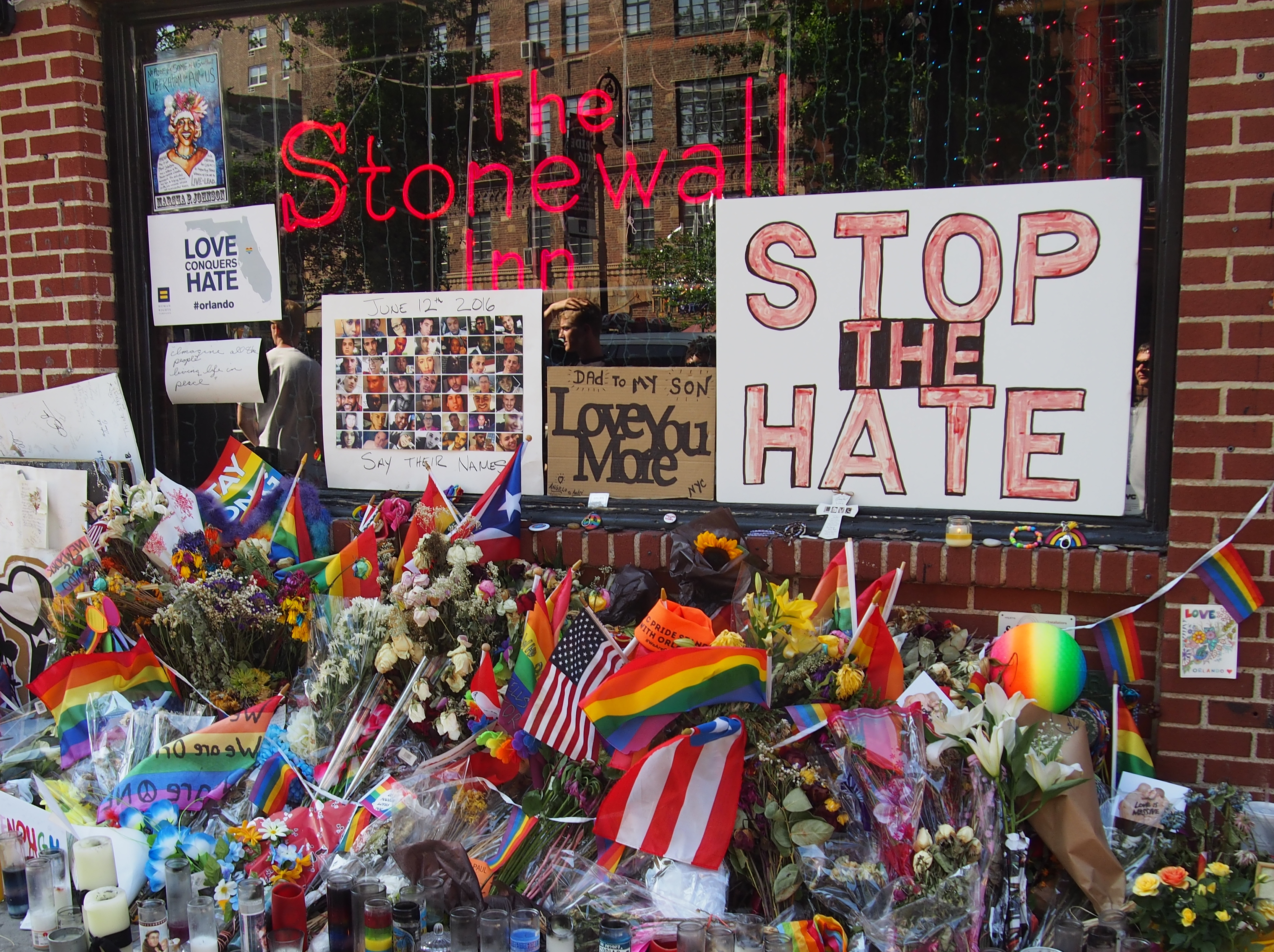 LGBTQ flags, flowers, and signs are laid on the ground outside the Stonewall Inn as a memorial. Some signs read, "Stop the hate" and "Love conquers hate".
