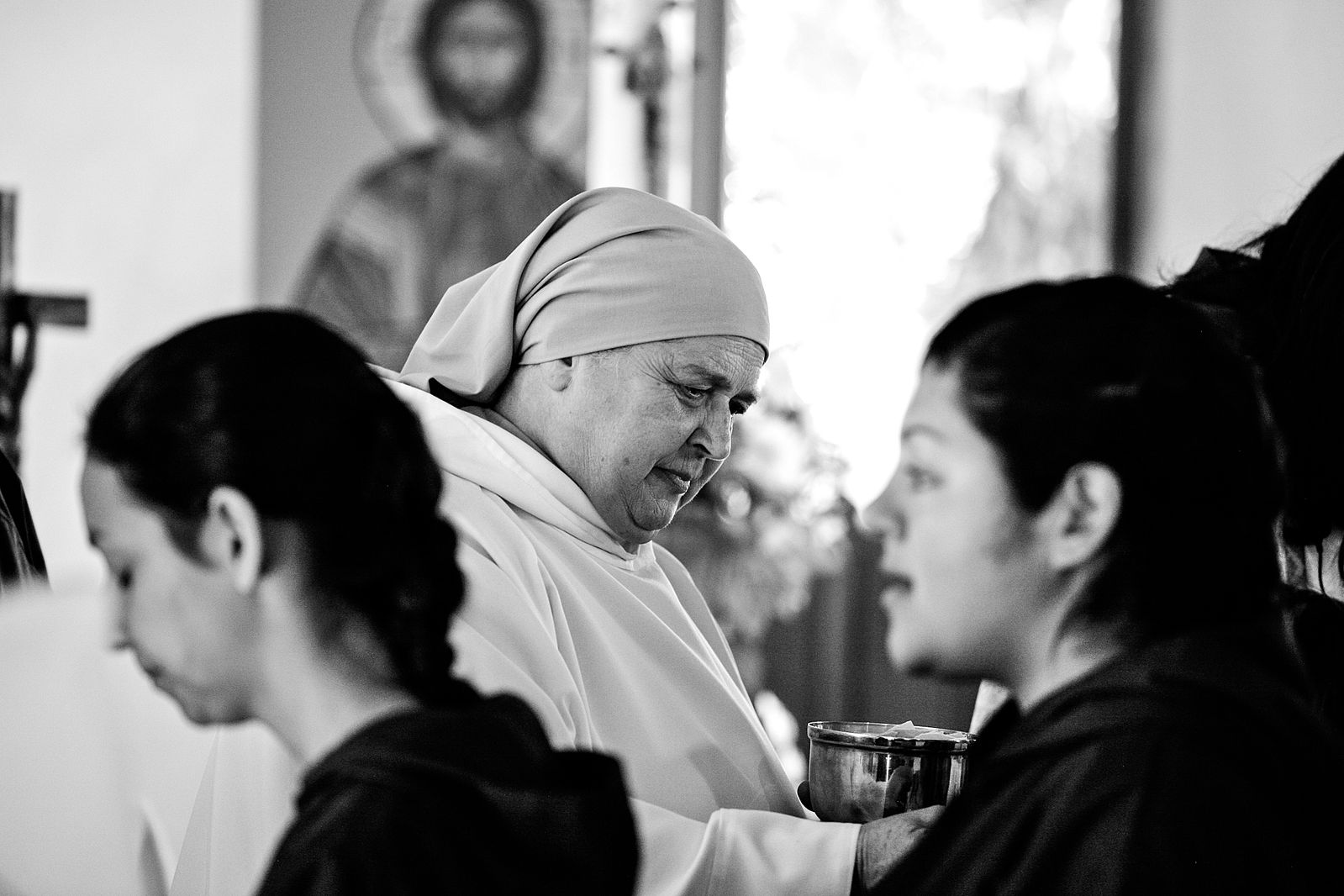 A woman with a head covering is in the middle of the photo holding a cup. There is a cross and a depiction of Jesus Christ out of focus in the background.
