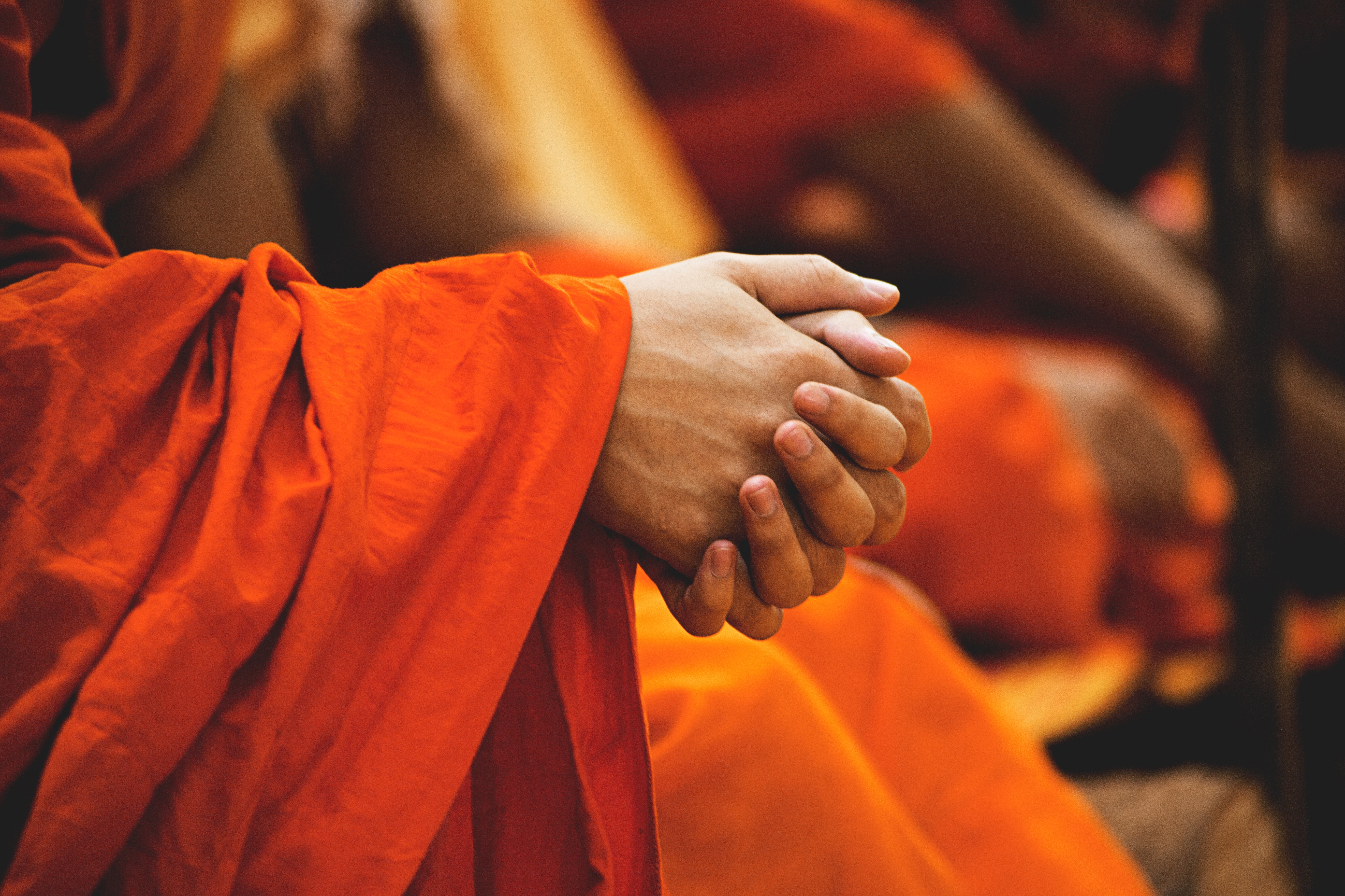 A person is sitting in the foreground but only their clasped hands and forearms are in the photo. They are wearing orange robes.