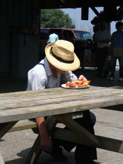 A young Mennonite boy in a straw hat is shown eating a piece of pizza at a picnic table.
