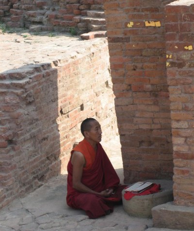 A man dressed in an orange robe is shown with his legs crossed, sitting within outdoor brick walls.