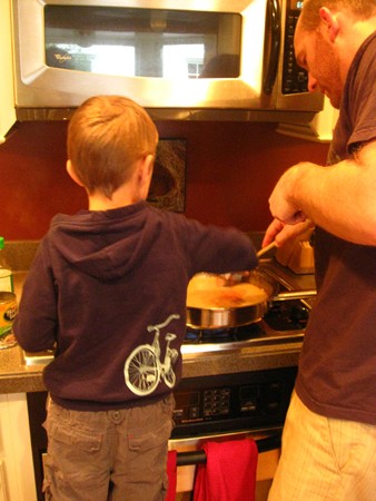 A man and young boy in the kitchen cooking at a stove.