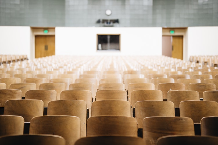 This photo is taken from the perspective of someone at the front of the lecture hall. In the foreground there are rows of wooden lecture hall chairs.
