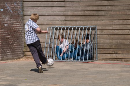 Boy kicking a soccer ball on a playground toward three other boys who are caged against a wall by a small metal goal post.
