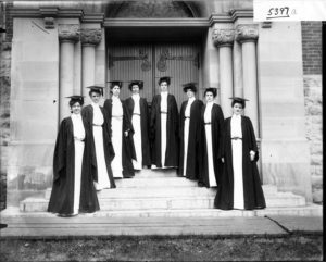 Eight women in dresses, caps, and gowns, standing on the steps of a college in a black in white photograph.