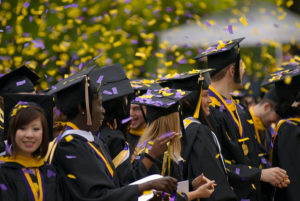 Graduating students in black caps and gowns, yellow tassels, with purple and gold confetti in the air.