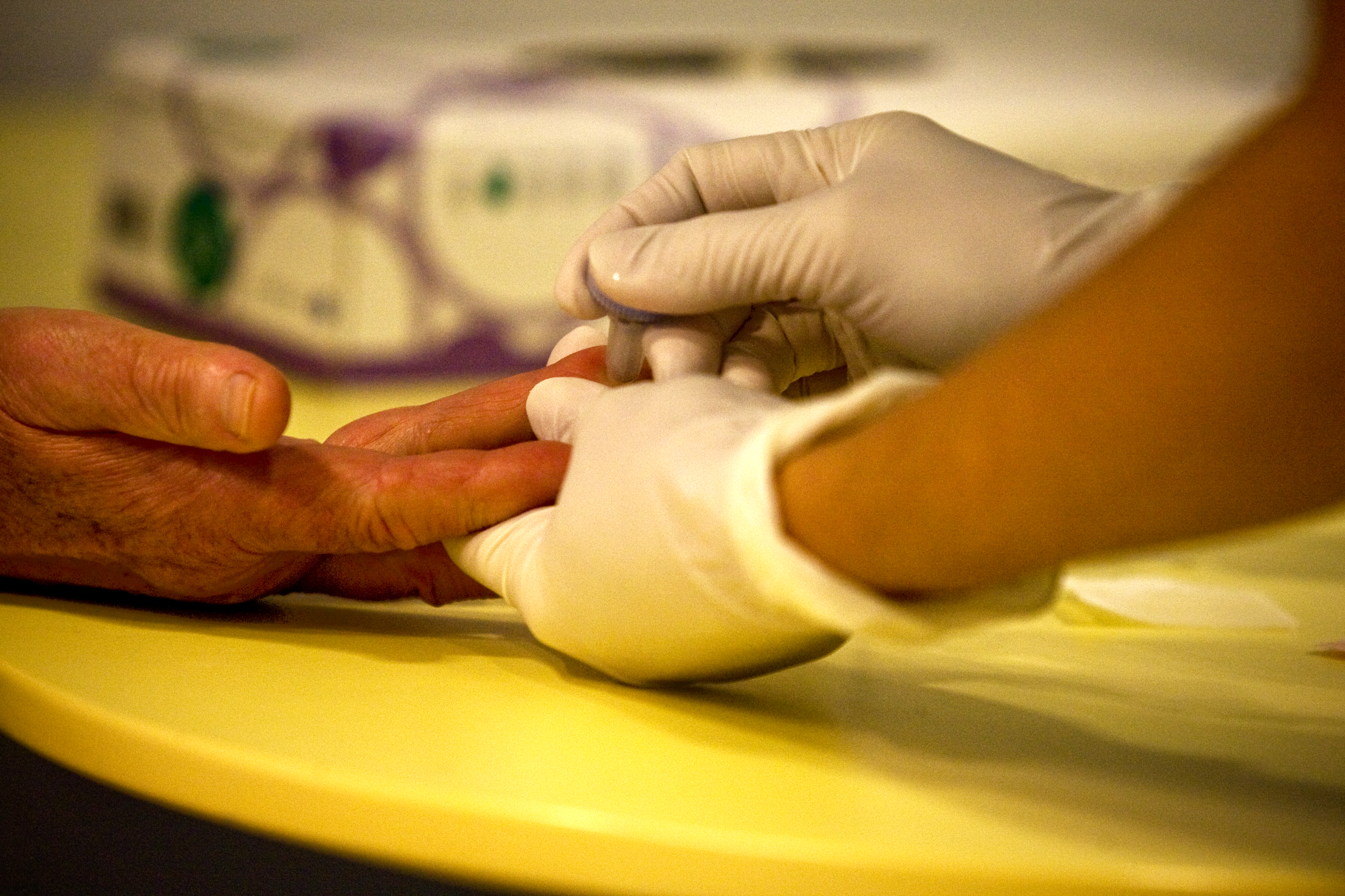 One person with latex gloves on is holding another persons hand while they use a medical device to prick the finger for blood.