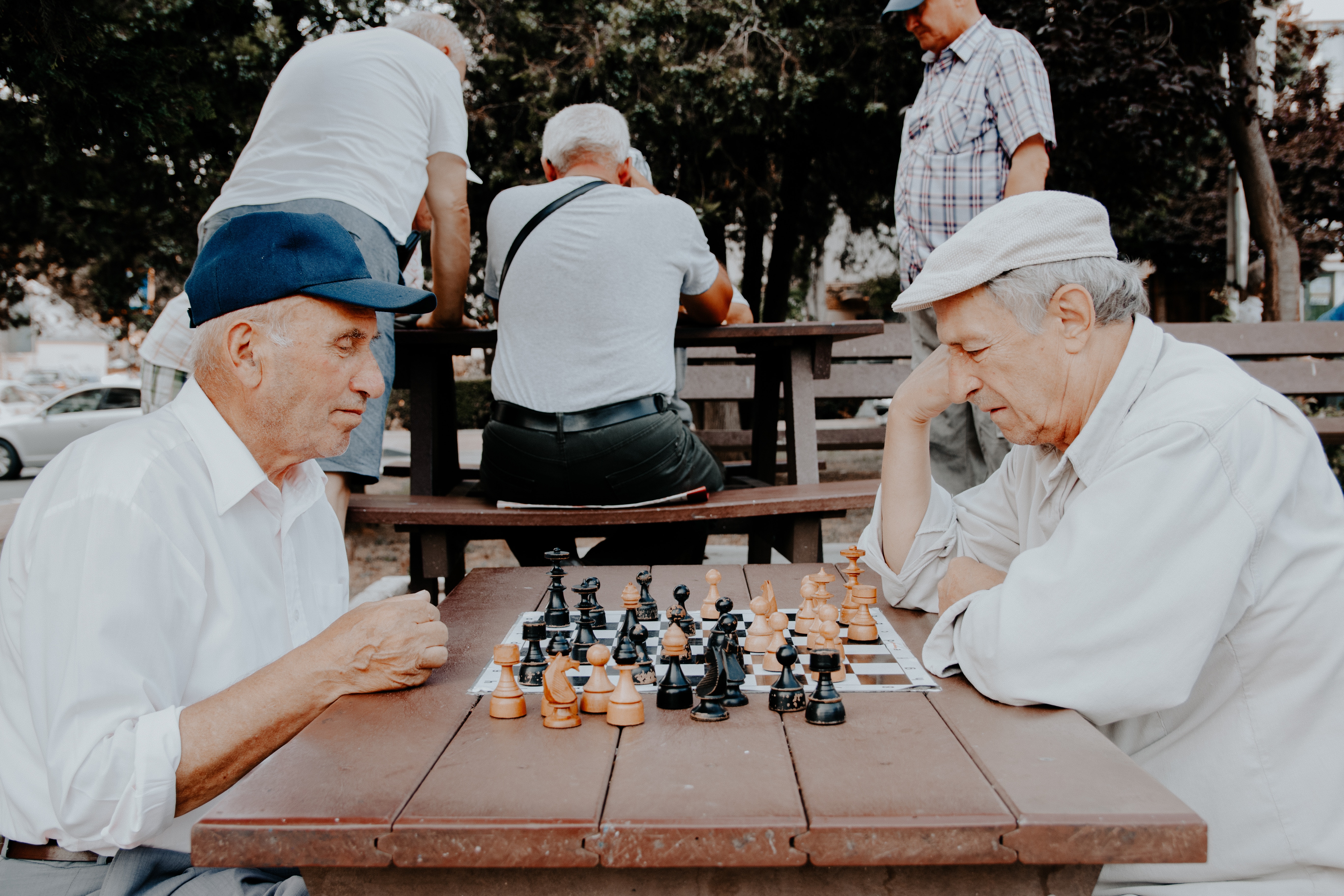 Two older men sit across from one another at a picnic table outside and are playing chess.