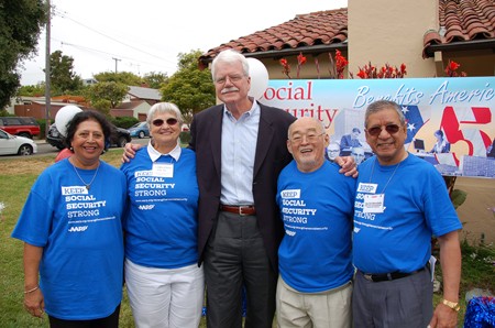 A tall man flanked by two elderly women on his right and two elderly men on his left. The elderly people are all wearing blue T-shirts that read, “Keep Social Security Strong: A A R P.” A banner in the background can also be seen, reading “Social Security Benefits America.”