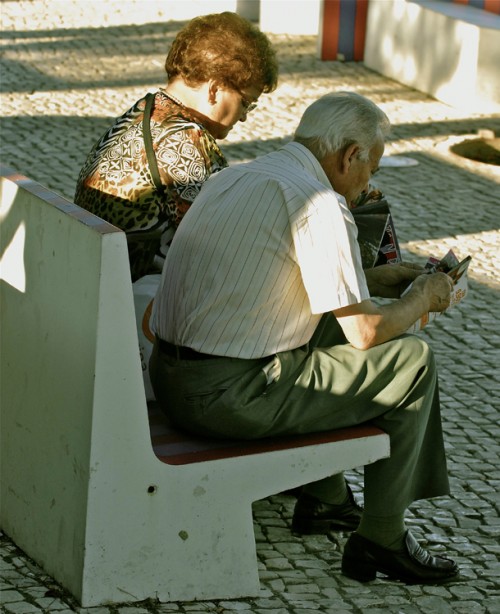 An elderly man and woman are shown sitting side by side on a bench.