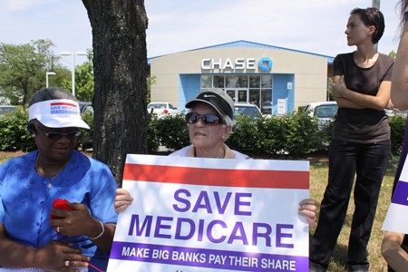 Two elderly women, one holding a red white and blue sign reading “Save Medicare: Make Big Banks Pay Their Share,” are shown sitting under some trees and in front of a suburban bank building.