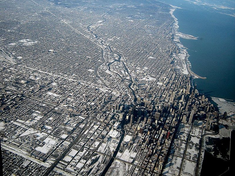 This photo is taken from high above Chicago and shows downtown Chicago and part of Lake Michigan. There are skyscrapers in the lower right of the photo and then a stretch of smaller buildings across the rest of the photo. Everything is very small and grey since these buildings are being photographed at a distance. Lake Michigan is on the upper right of the photo and is blue green.