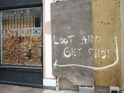 A photo of a damaged storefront from Hurricane Katrina with a sign posted on the wall that reads, "Loot and get shot"