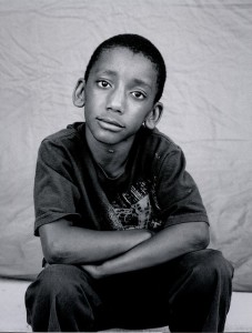 boy looking reflectively at the camera for his picture, while he sits with his arms crossed in front of him on his knees.