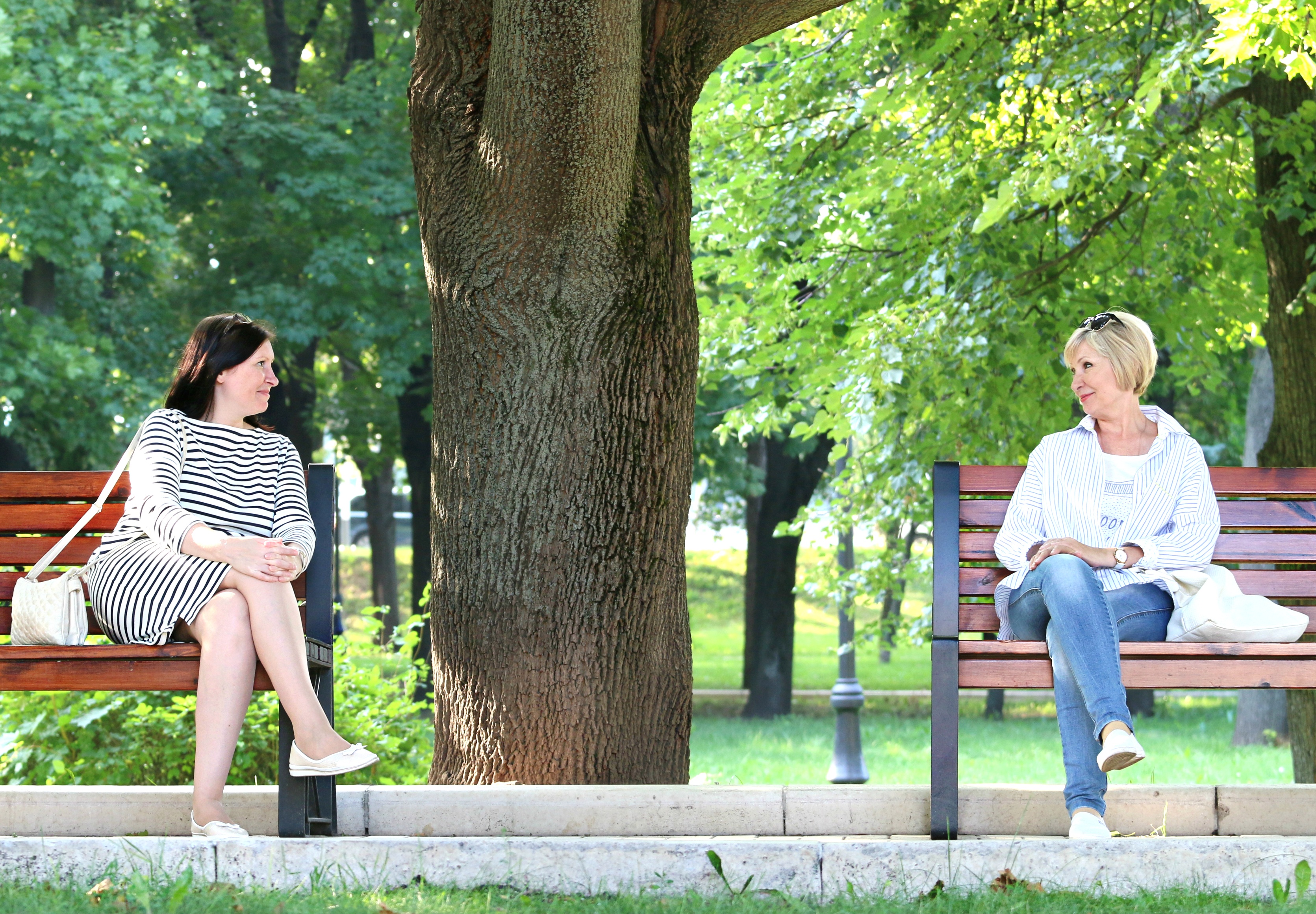 Two women sitting on different park benches are smiling at each other
