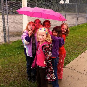 Un grupo de chicas jóvenes se paran juntas bajo una sombrilla en el patio de recreo.