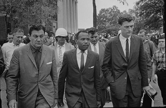 A Black man wearing a suit escorted by other men wearing suits. James Meredith, accompanied by U.S. Marshalls, walks to class at the University of Mississippi in 1962. Meredith was the first African-American student admitted to the still segregated Ole Miss.