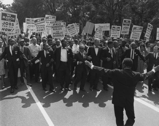 A crowd of people wearing suits or formal dress, mostly Black but a few White. They hold signs asking for jobs, the end of segregation, and equal rights for all.