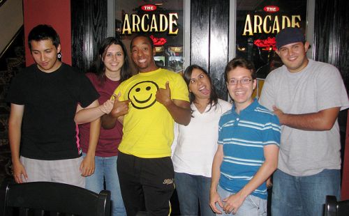 A diverse group of older teenagers (four boys and two girls), smile and make silly faces while standing in front of the arcade.