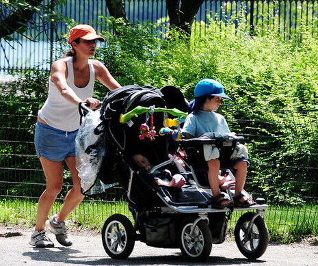 A woman jogs through a park with three young children in a stroller.