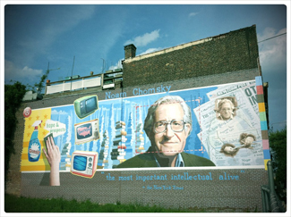 Una fotografía muestra un mural en el costado de un edificio. El mural incluye la cara de Chomsky, junto con algunos periódicos, televisores y productos de limpieza. En la parte superior del mural, se lee “Noam Chomsky”. En la parte inferior del mural, se lee “el intelectual más importante vivo”.