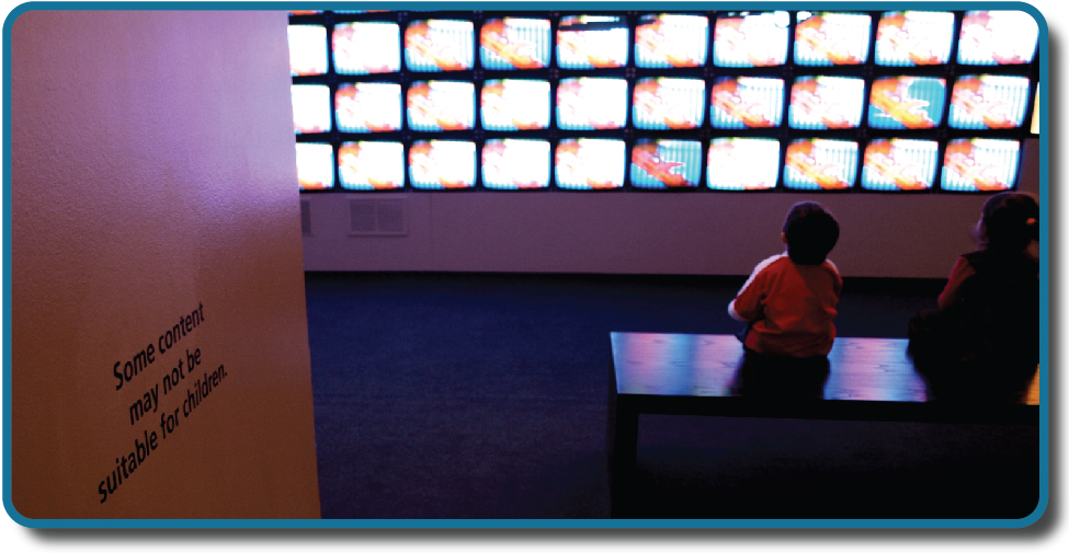 As crianças se sentam em frente a um banco de telas de televisão. Uma placa na parede diz: “Alguns conteúdos podem não ser adequados para crianças”.
