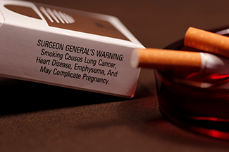 Uma fotografia mostra um maço de cigarros e cigarros em um cinzeiro. O maço de cigarros diz: “Aviso do cirurgião geral: fumar causa câncer de pulmão, doenças cardíacas, enfisema e pode complicar a gravidez”.