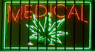 Una fotografía muestra una ventana con un letrero de neón. El letrero incluye la palabra “médico” sobre la forma de una hoja de marihuana.
