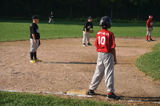 Uma fotografia de crianças jogando beisebol é mostrada. Cinco crianças estão na foto, duas em uma equipe e três na outra.