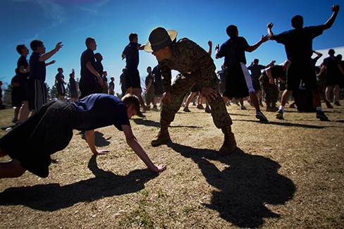 Una fotografía muestra a una persona haciendo pushups mientras un líder militar se para sobre la persona; otras personas están haciendo saltos de fondo.