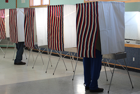 Uma fotografia mostra uma fileira de cabines de votação com cortinas; duas são ocupadas por pessoas.