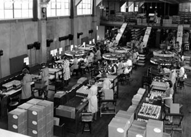 Uma fotografia mostra um armazém cheio de pessoas trabalhando com máquinas ao longo das linhas de montagem.