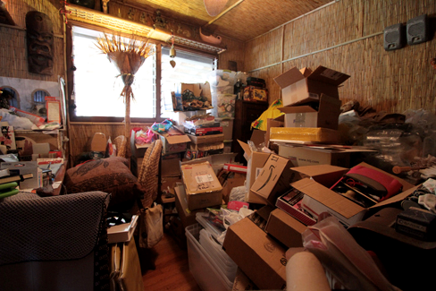 Uma fotografia mostra uma pequena sala contendo grandes pilhas de caixas, repletas de papéis, fichários e vários outros pertences. Muitos dos móveis e do piso estão escondidos sob esses outros objetos.