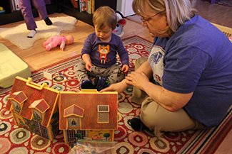 Un adulto y un niño pequeño son representados sentados en una alfombra junto a una casa de juguetes.