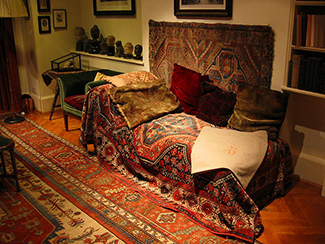 Esta fotografia mostra a aparência do famoso sofá psicanalítico de Freud. O sofá está coberto de tapeçarias e travesseiros, e o quarto é decorado com esculturas, livros e quadros na parede.