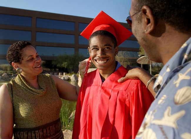 Padres sonrientes se paran con su hijo adulto joven que está vestido con una gorra y un vestido de graduación.” title="Padres sonrientes se paran con su hijo adulto joven que está vestido con una gorra y bata de graduación.