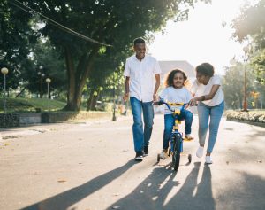 A father and mother walk alongside their daughter who is learning to ride a bike