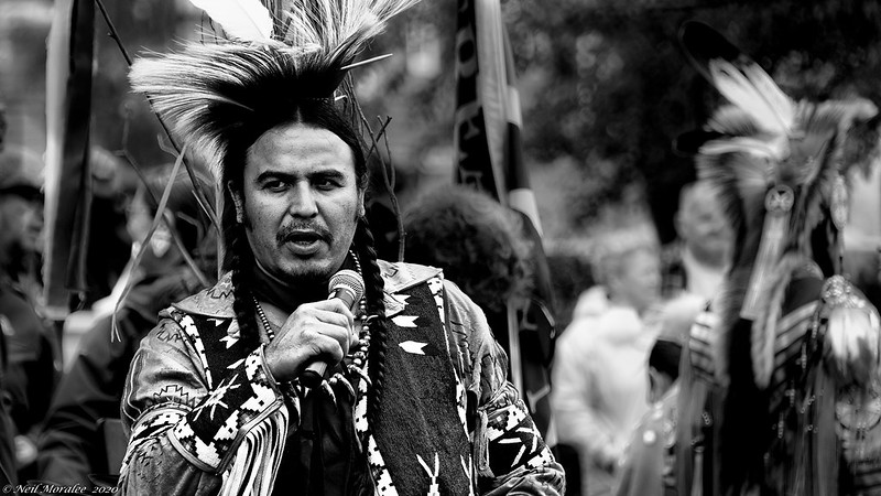 man wearing traditional Native American clothing speaks into a microphone