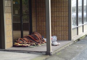 Photo of a person with a blanket, sleeping in the front of a building.