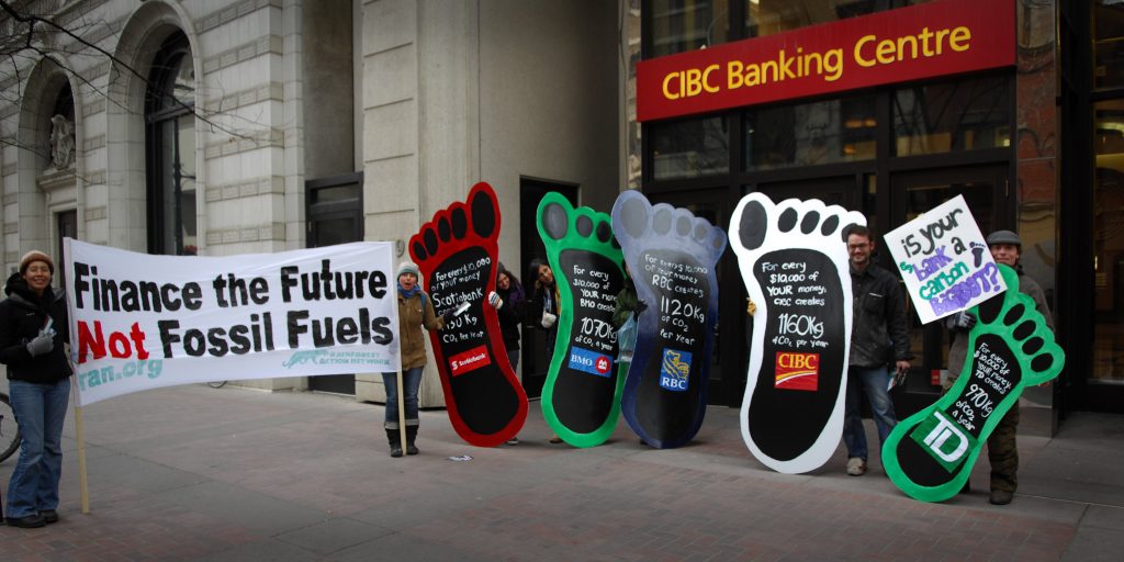 Photograph of people holding posters of large feet in front of a bank protesting.