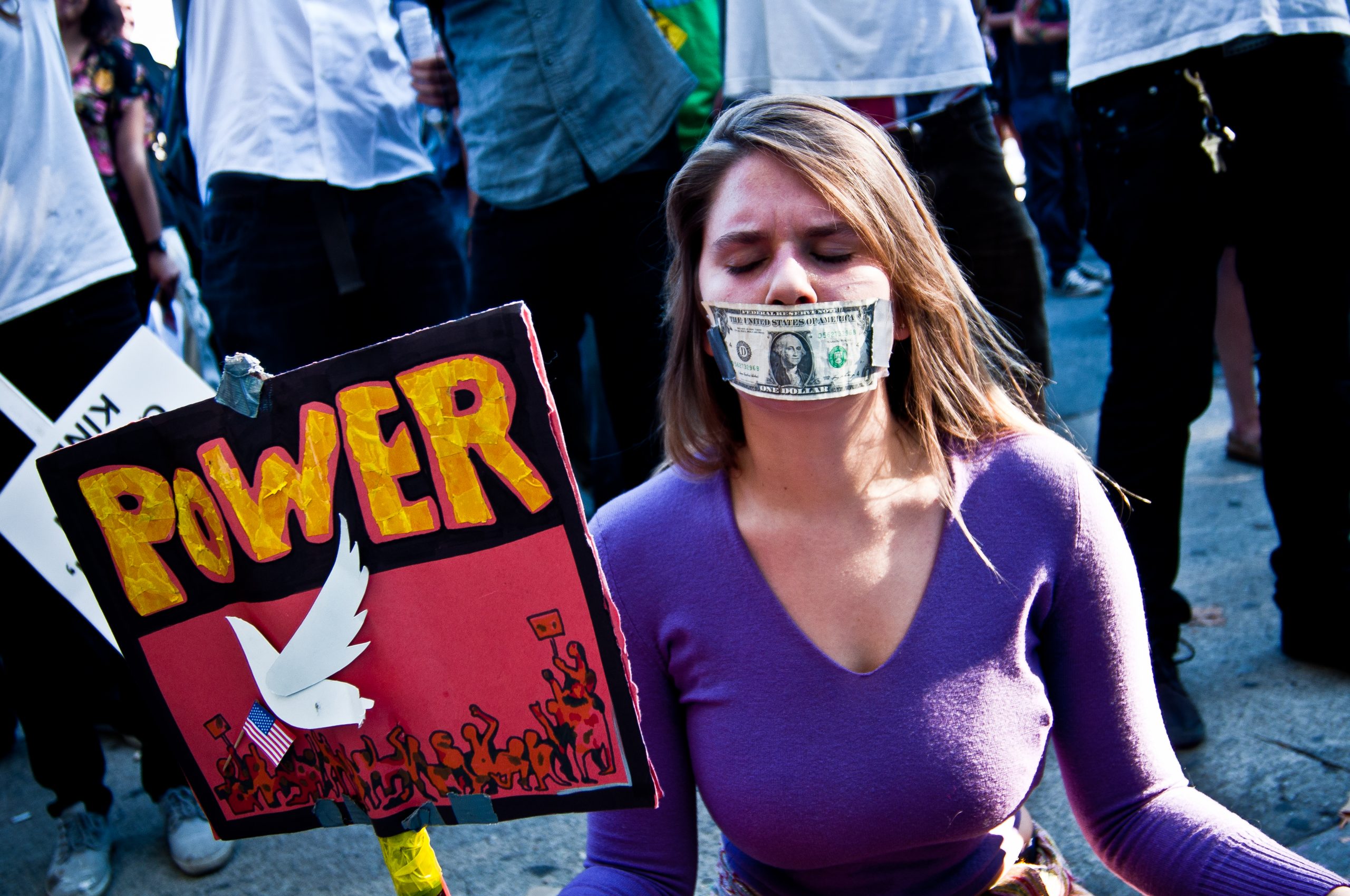 Photograph of a woman with a sign saying POWER. She has a dollar bill taped to her mouth.
