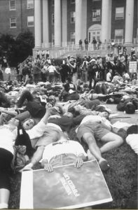 Black and white photo of thousands of demonstrators lying down on the lawn, surrounded by police on horseback, in front of a building with pillars.