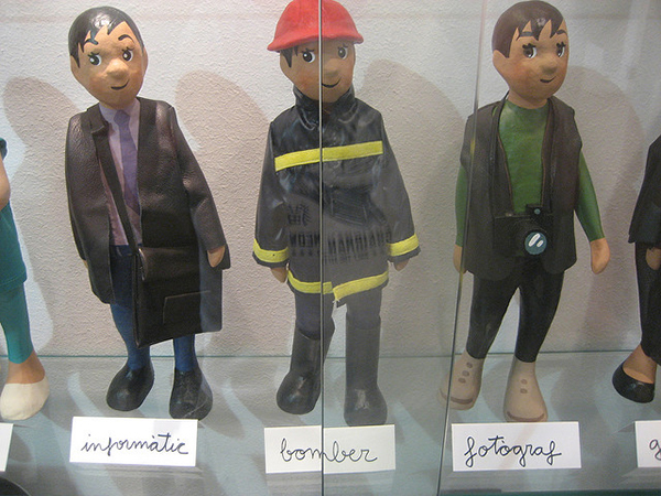 A series of the same papier mache doll dressed as different occupations. They are labeled: Informatic (suit), Bomber (fire fighter), and Fotograph(camera around neck)