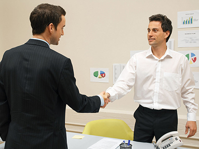 Two men shaking hands over a table, one is in a suit and the other is in a dress shirt and slacks.