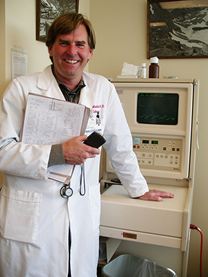 Smiling doctor in a white coat smiling in front of a piece of medical equipment. He holds a chart and cellphone in his arm.