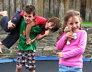 Three children play on a trampoline. Two boys are posed growling in a wrestling hold and a girl is smiling and snuggling a stuffed animal.