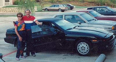 Older couple smiling and posing in front of a shiny black sports car.