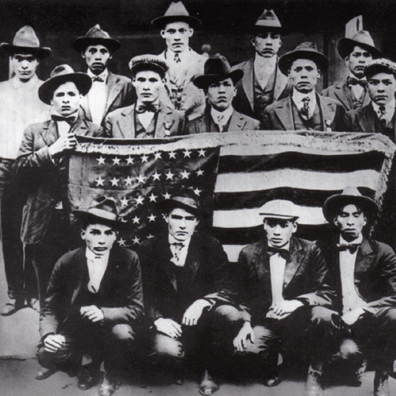 A group of Choctaw men pose around a large American flag in civilian dress