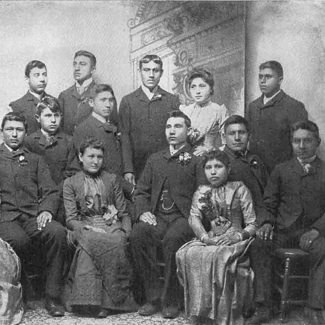 AMerican Indian students in white American clothing and hairstyles. Caption reads Educating the Indian Race. Graduating Class of Carlisle, PA.