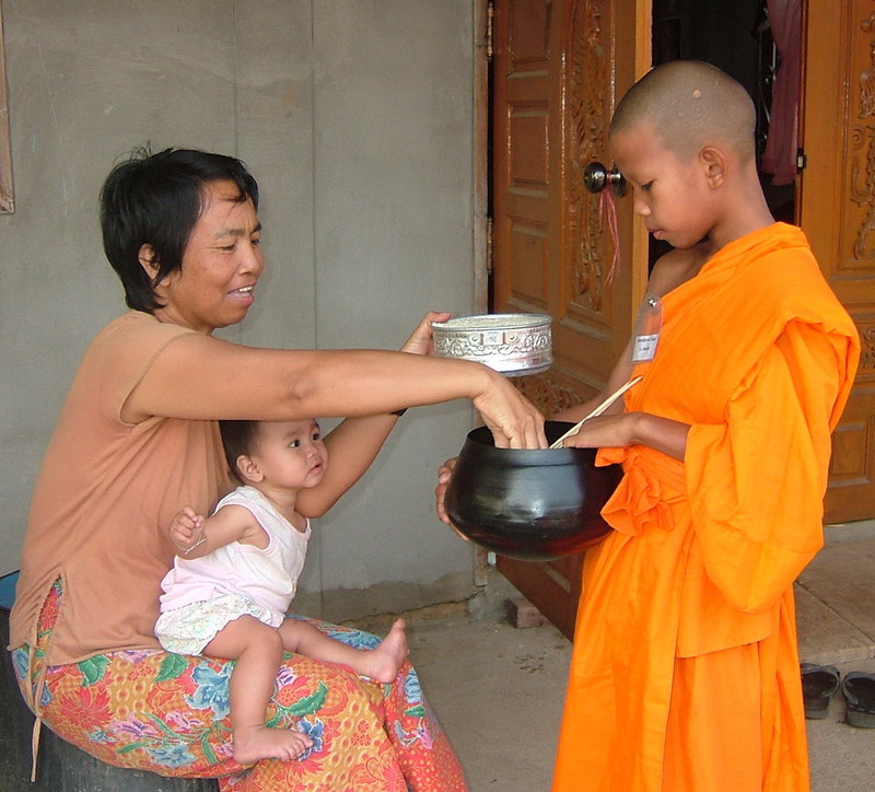 A Buddhist woman with a baby on her lap places food into the alms bowl of a young Buddhist priest dressed in traditional orange robes. 